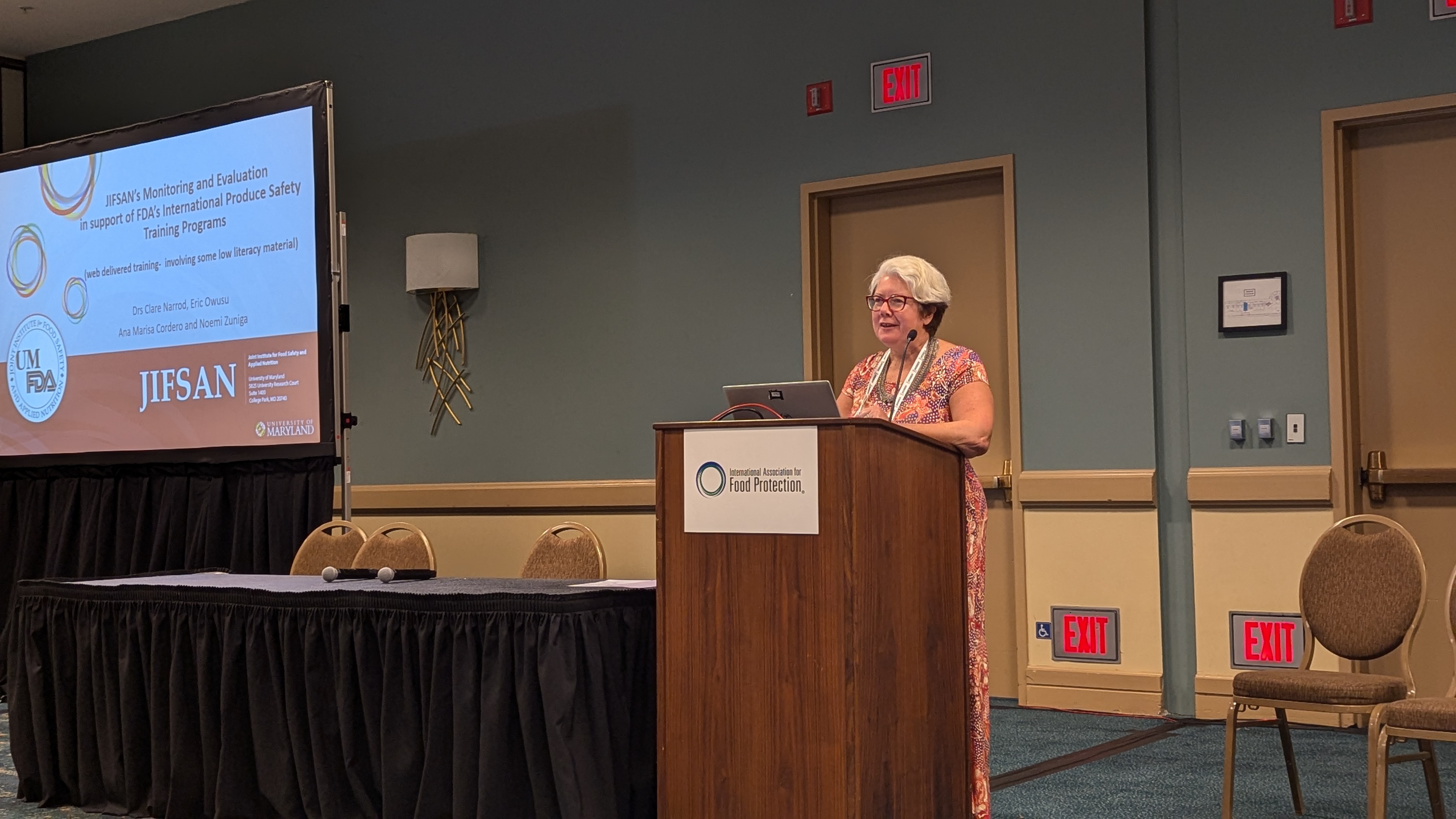 Dr. Clare Narrod stands at a lectern giving a presentation titled JIFSAN Monitoring and Evaluation in support of FDA's International Produce Safety Training Programs