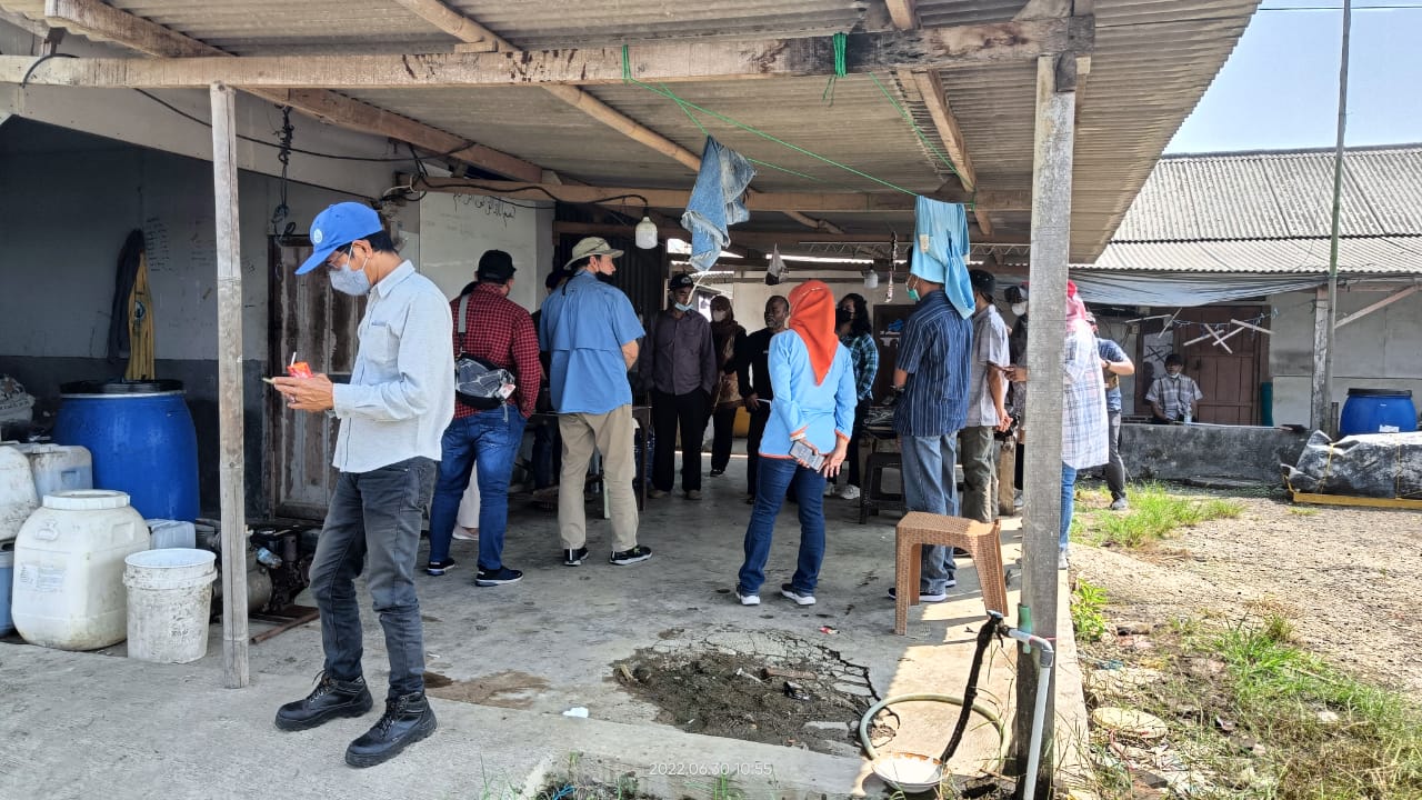 Good Aquaculture Practices students stand under a timber-framed awning at a shrimp farm in Indonesia. Blue and white plastic drums are seen to the side, and a garden hose connected to PVC pipe is visible in the foreground.