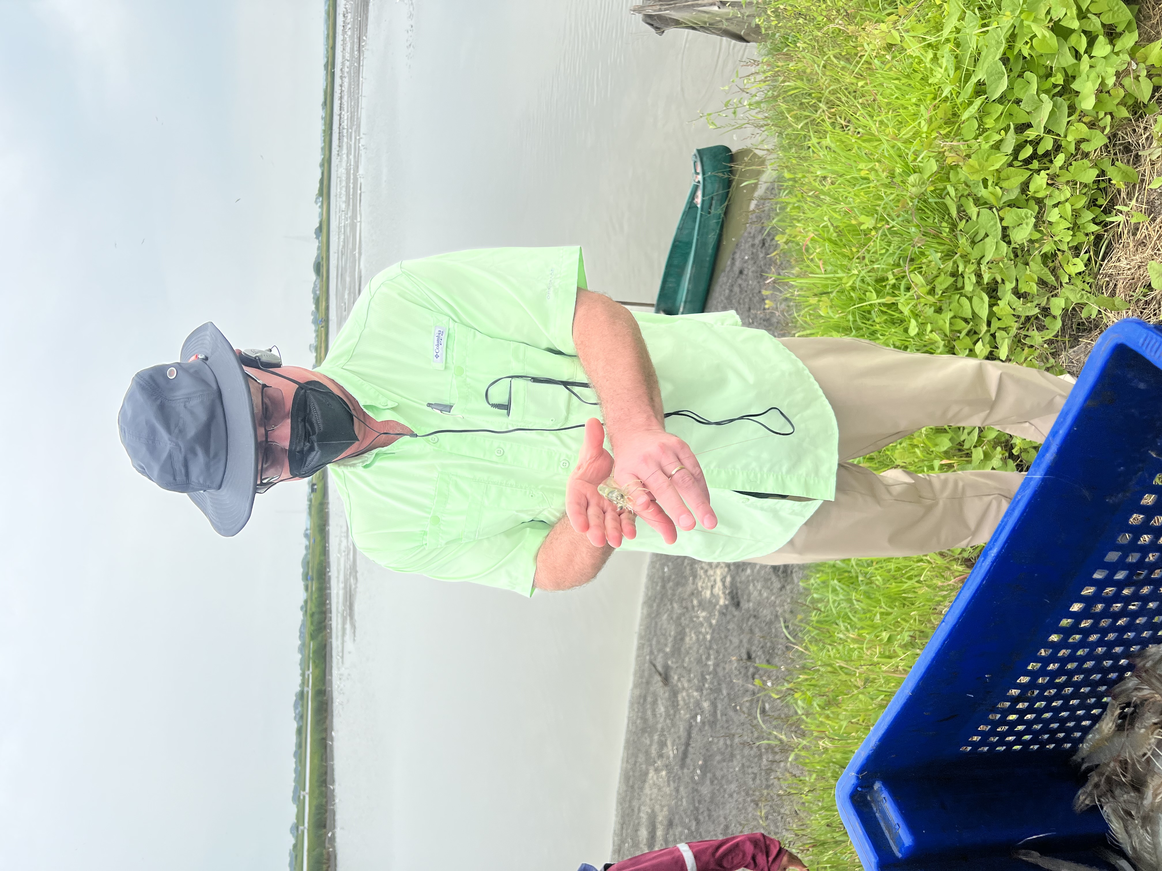 Michael Schwarz is seen holding a shrimp. A body of water with a small boat is in the background, and a basket full of shrimp is visible in the foreground.