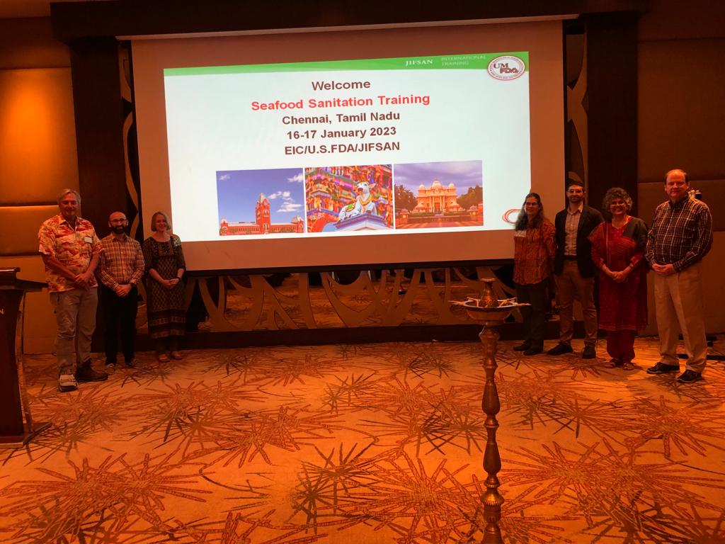 Seven conference participants stand in front of a projected slide introducing the Seafood Sanitation Training session. A tall South Indian lamp is in the foreground.