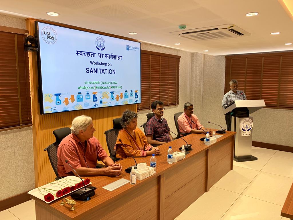 A man speaks at a lectern while four conference participants sit at a table in front of a slide introducing the sanitation workshop in English and Hindi.